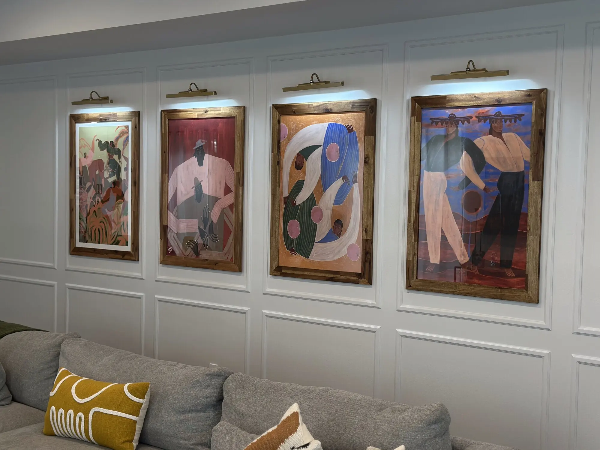 Four large framed art panels on a panelled wall, each lit by its own picture light